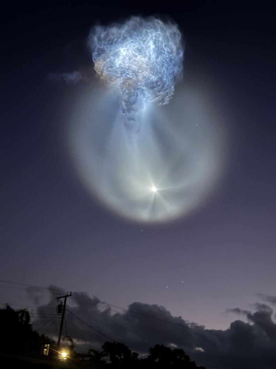 Blue-white rocket plume and a pale circular glow high above silhouetted trees, power lines and clouds at dawn