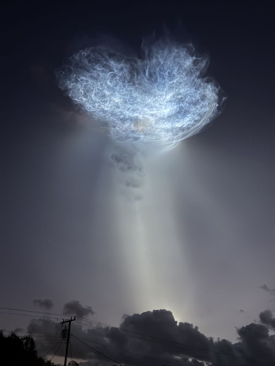 Wide, feathery white rocket exhaust cloud with a pale beam of light below it, above dark clouds and a utility pole