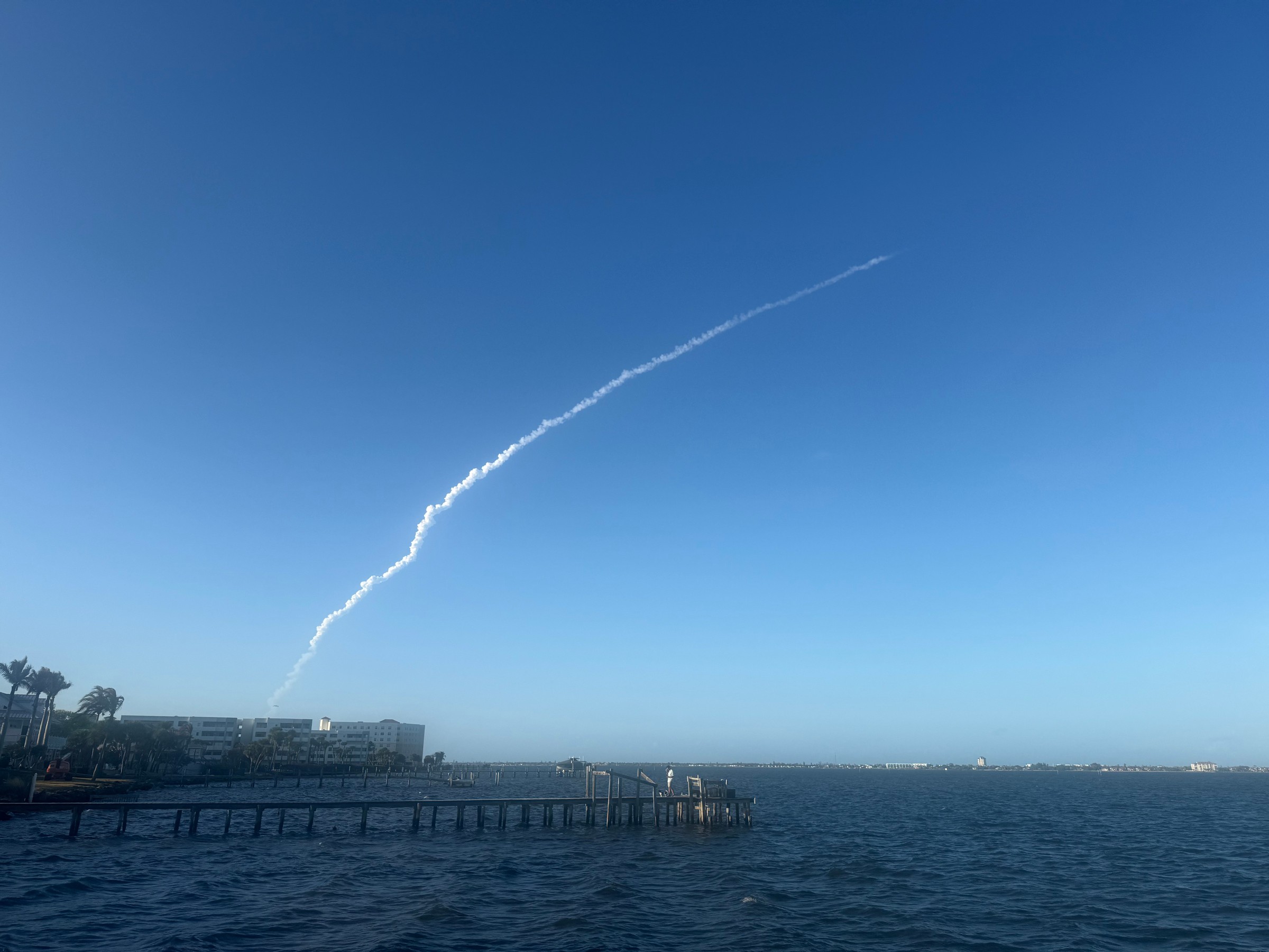 A thin white rocket exhaust trail arcs up into a clear blue evening sky above the Indian River, with a long wooden dock and condo buildings at the waterline