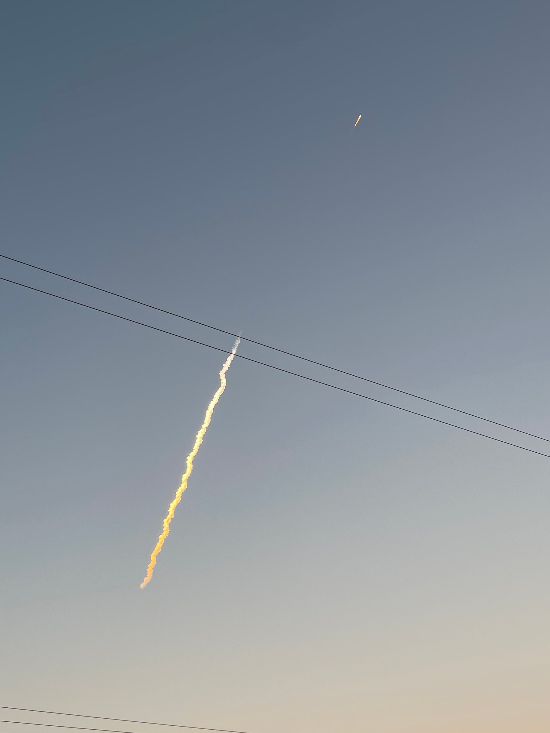 A golden, sunlit rocket plume rises diagonally through a blue-gray twilight sky, with a bright point of engine light above it and power lines crossing the frame