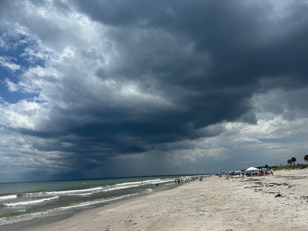 A towering dark storm cloud drops a gray rain shaft over the ocean beyond a crowded beach with tents and umbrellas