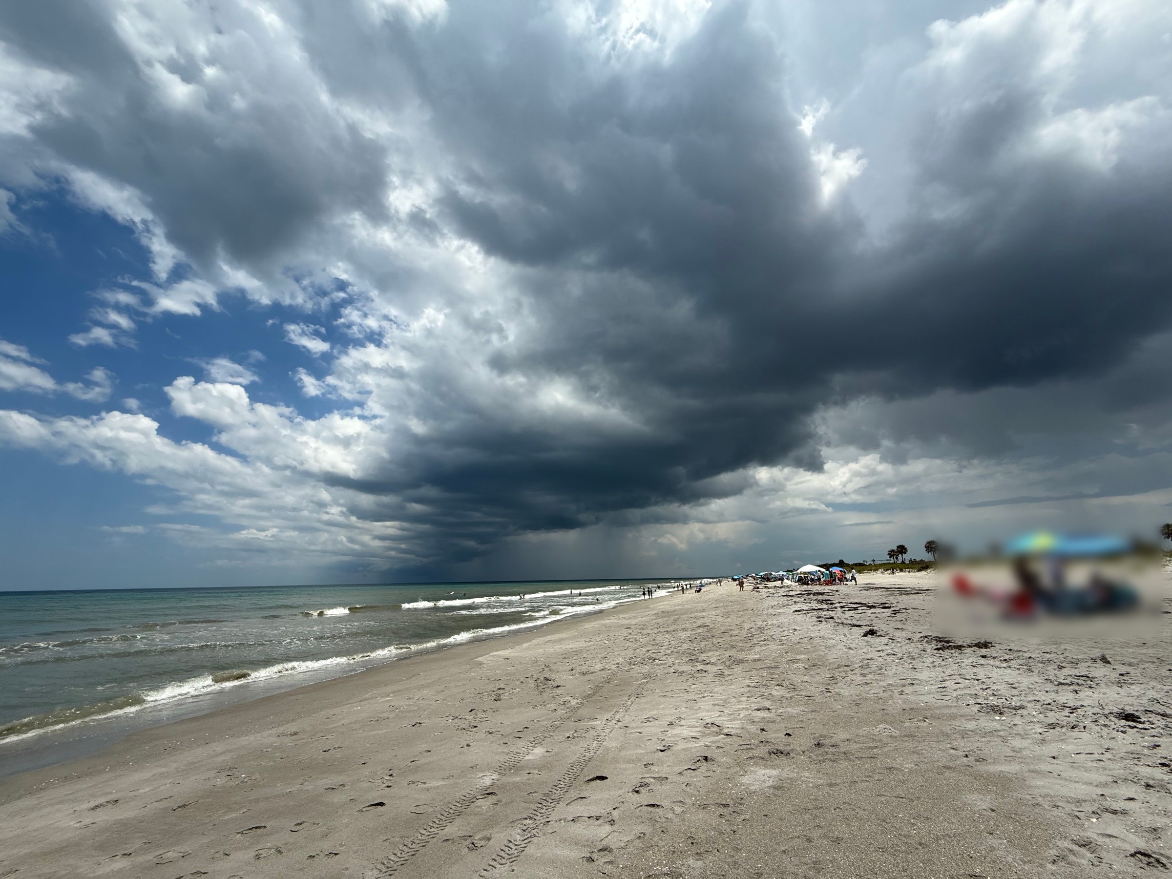 A storm cell with a visible rain shaft hangs over the Atlantic off a wide sandy beach under partly blue skies