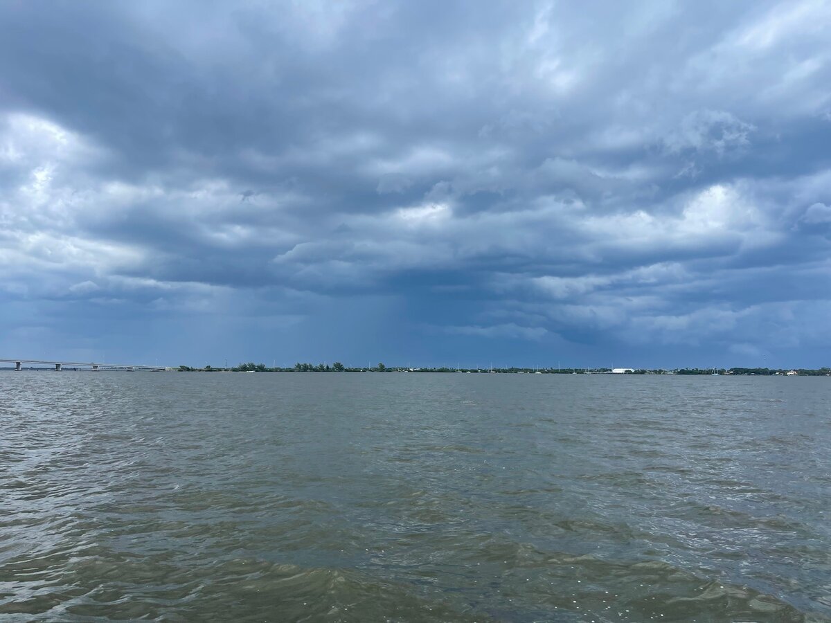 Heavy, layered storm clouds and faint rain hang low over choppy gray-green lagoon water and a distant shoreline