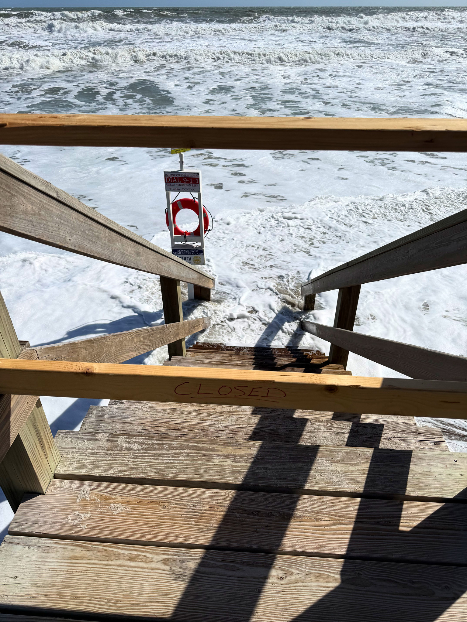 Wooden beach-access stairs with "CLOSED" handwritten on the rail and a life-ring station beside them, with foamy surf covering the beach up to the bottom step