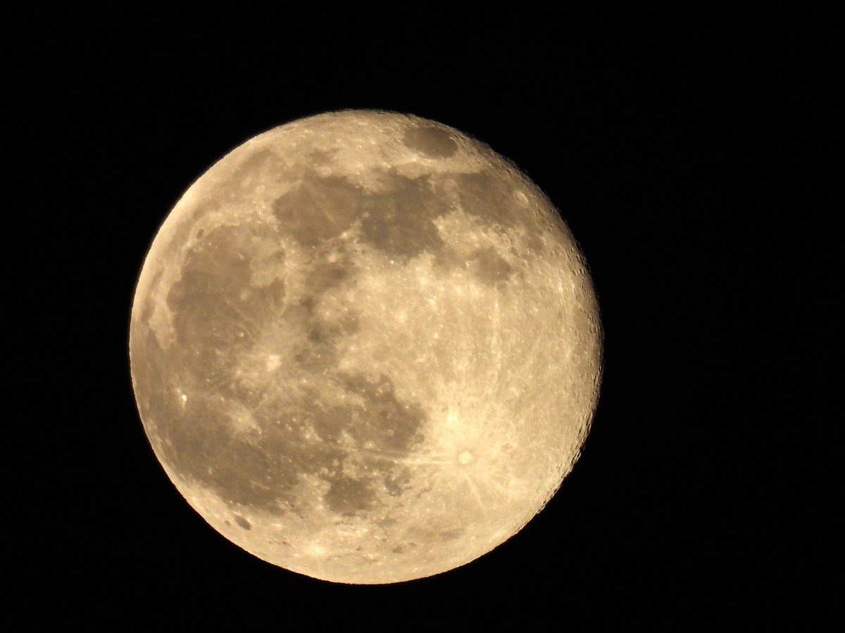 A warm-toned nearly full moon with dark lunar seas and rayed craters against a black sky