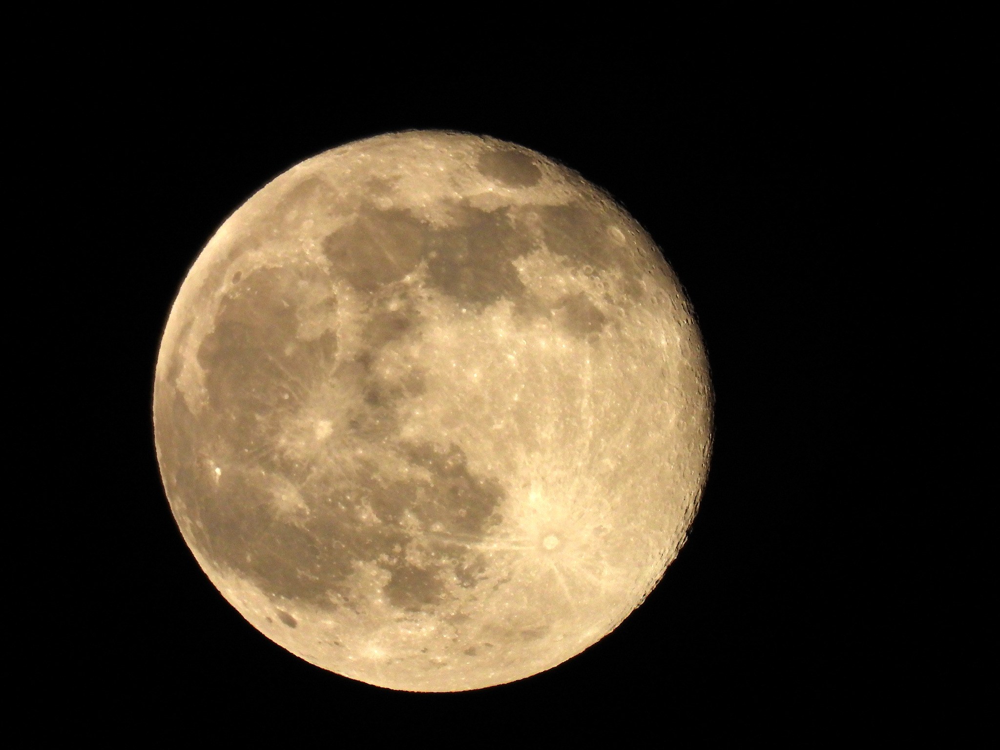 A warm-toned nearly full moon with dark lunar seas and rayed craters against a black sky