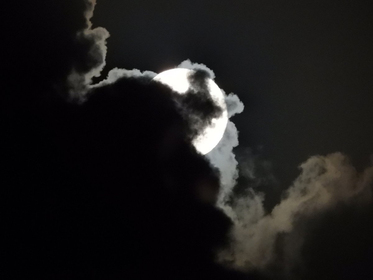 The bright moon peeks out from behind a dark, billowing cloud whose edges glow silver in the moonlight