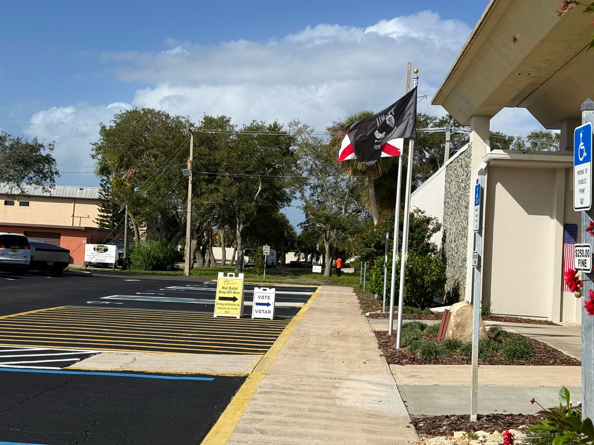 A yellow Mail Ballot Drop-Off Here sign and a VOTE/VOTAR sign with arrows stand beside a walkway next to Satellite Beach City Hall