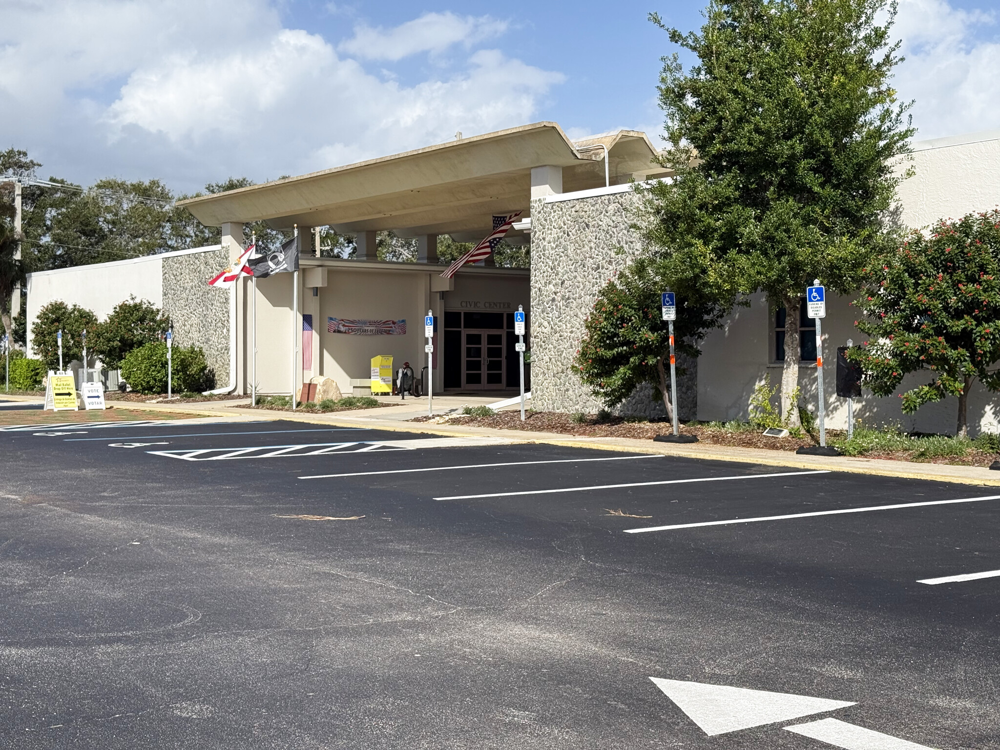 Front of Satellite Beach City Hall, a low stone-and-stucco building lettered Civic Center, with flags, a yellow sign by the entrance and voting signs at the edge of the parking lot