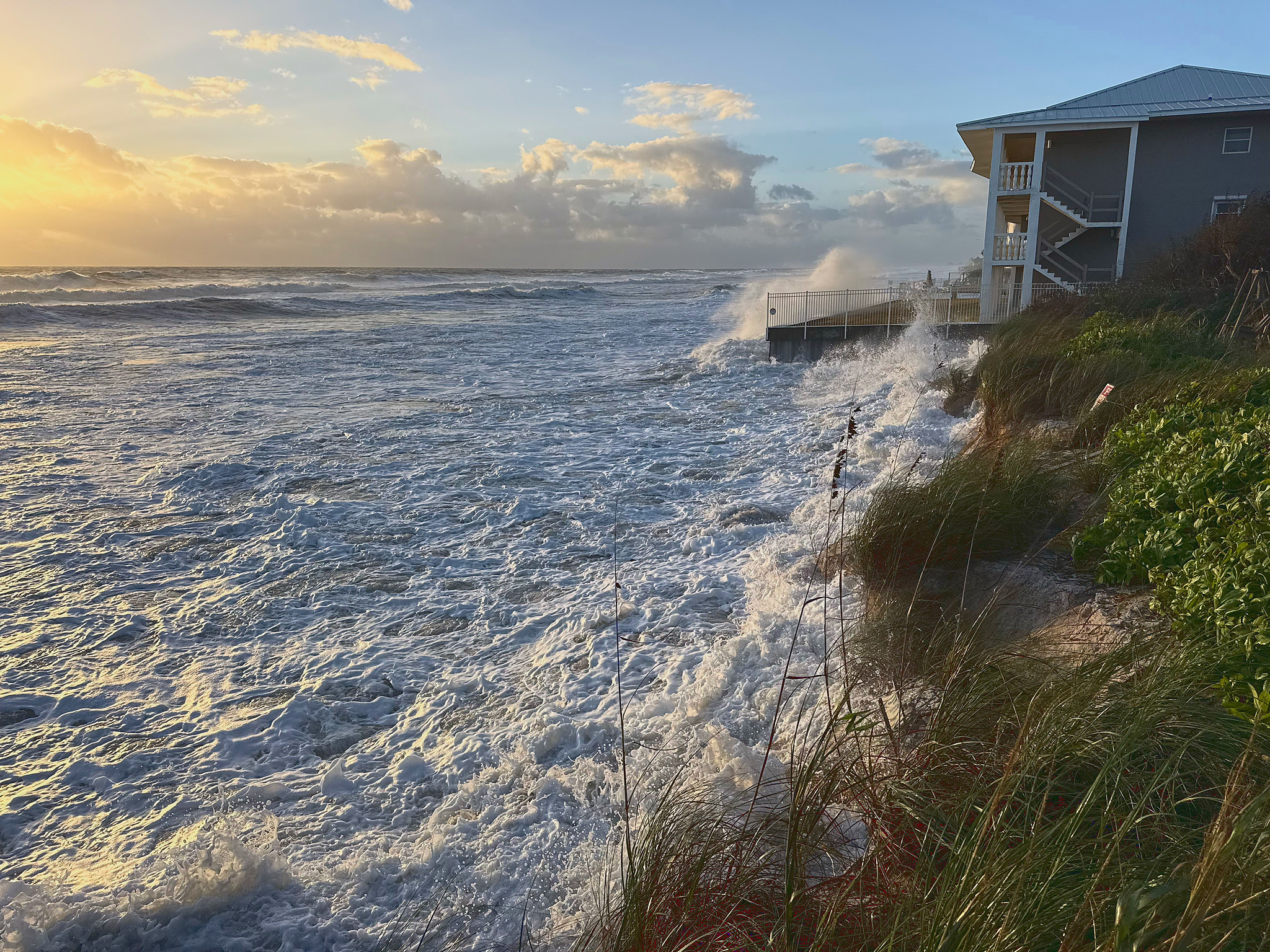 Foamy surf breaks into dune grass and sprays over a seawall corner beside a gray two-story beach house, under a partly cloudy sky