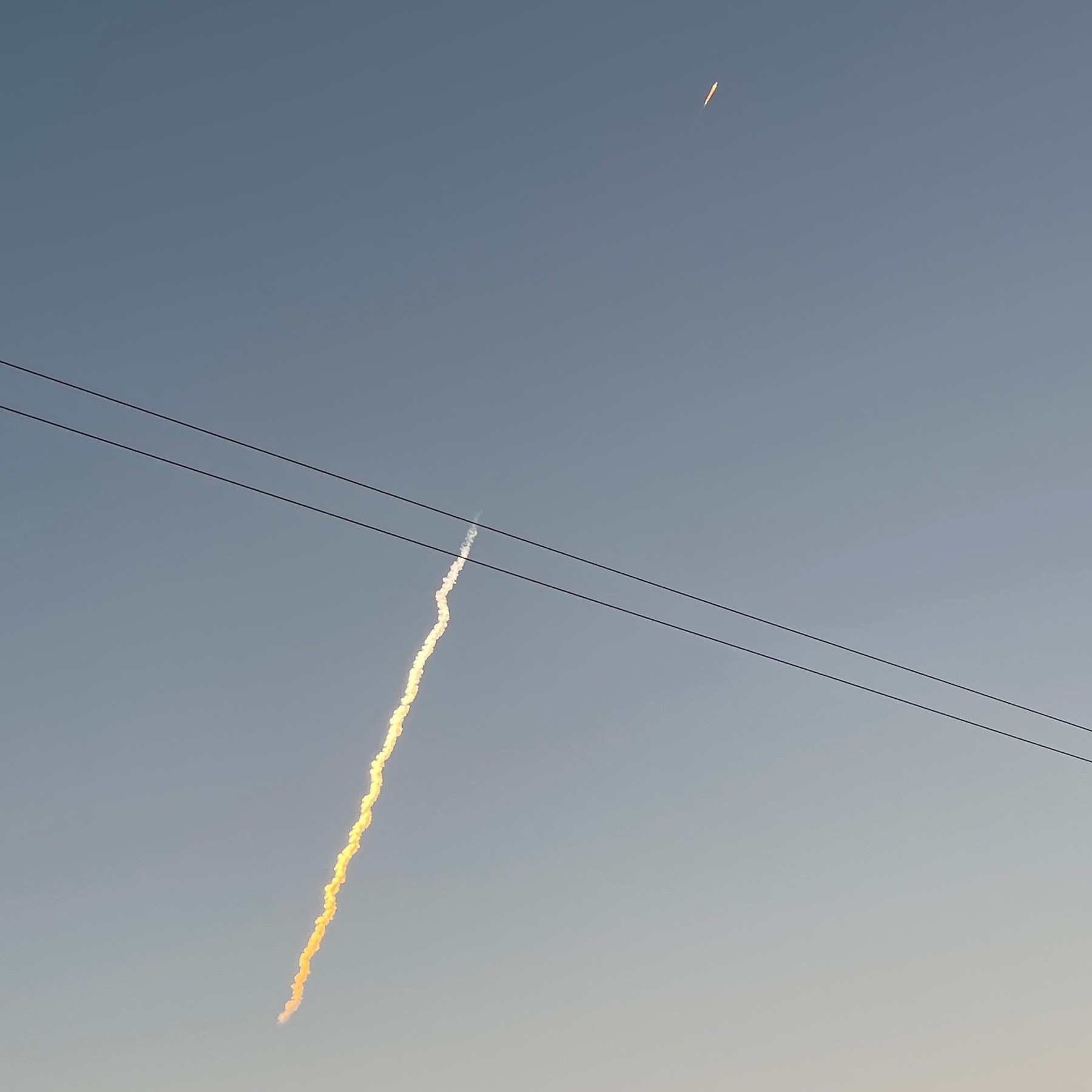 A sunlit Falcon 9 launch plume in the twilight sky, with the rocket's engine glow above it