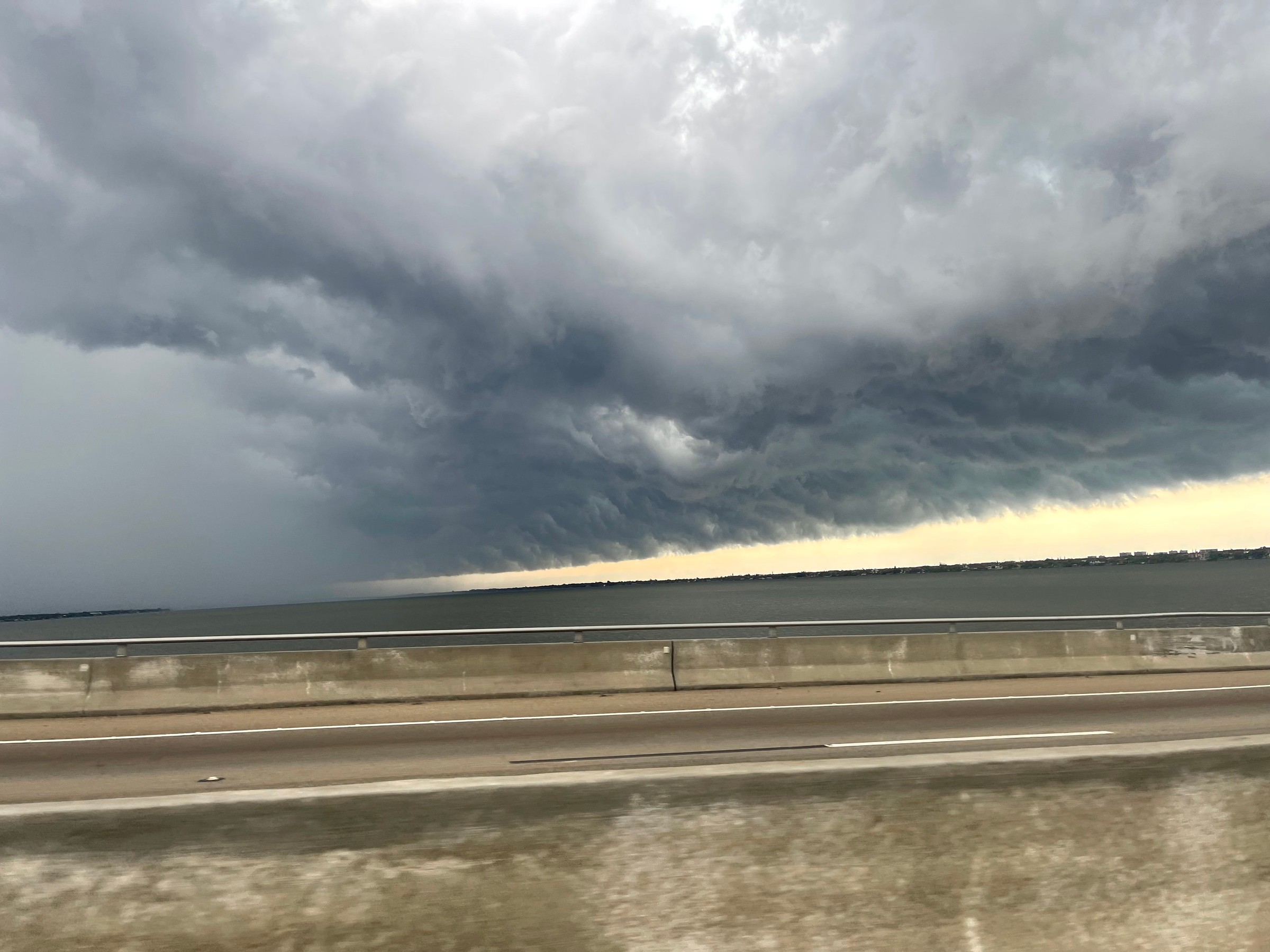A massive dark gray shelf cloud with a churning underside sweeps over the Indian River, with a band of pale yellow light beneath it and a curtain of rain at left, seen past a concrete causeway barrier