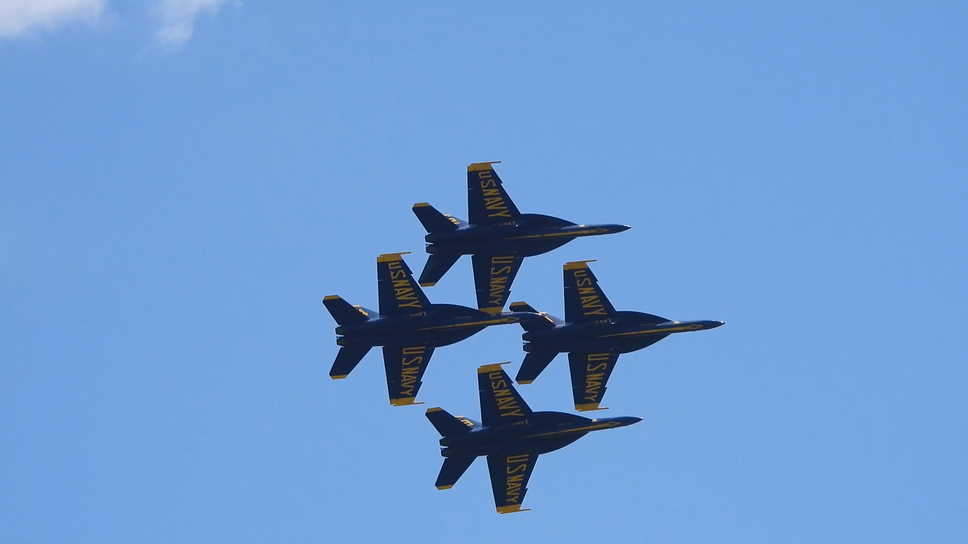Four blue-and-gold Navy jets fly in a tight diamond formation, seen from below against a blue sky