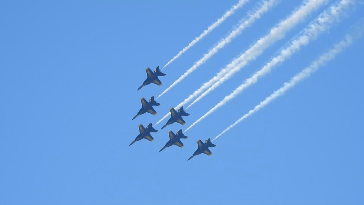Six jets fly in close formation, trailing long parallel white smoke lines across a blue sky