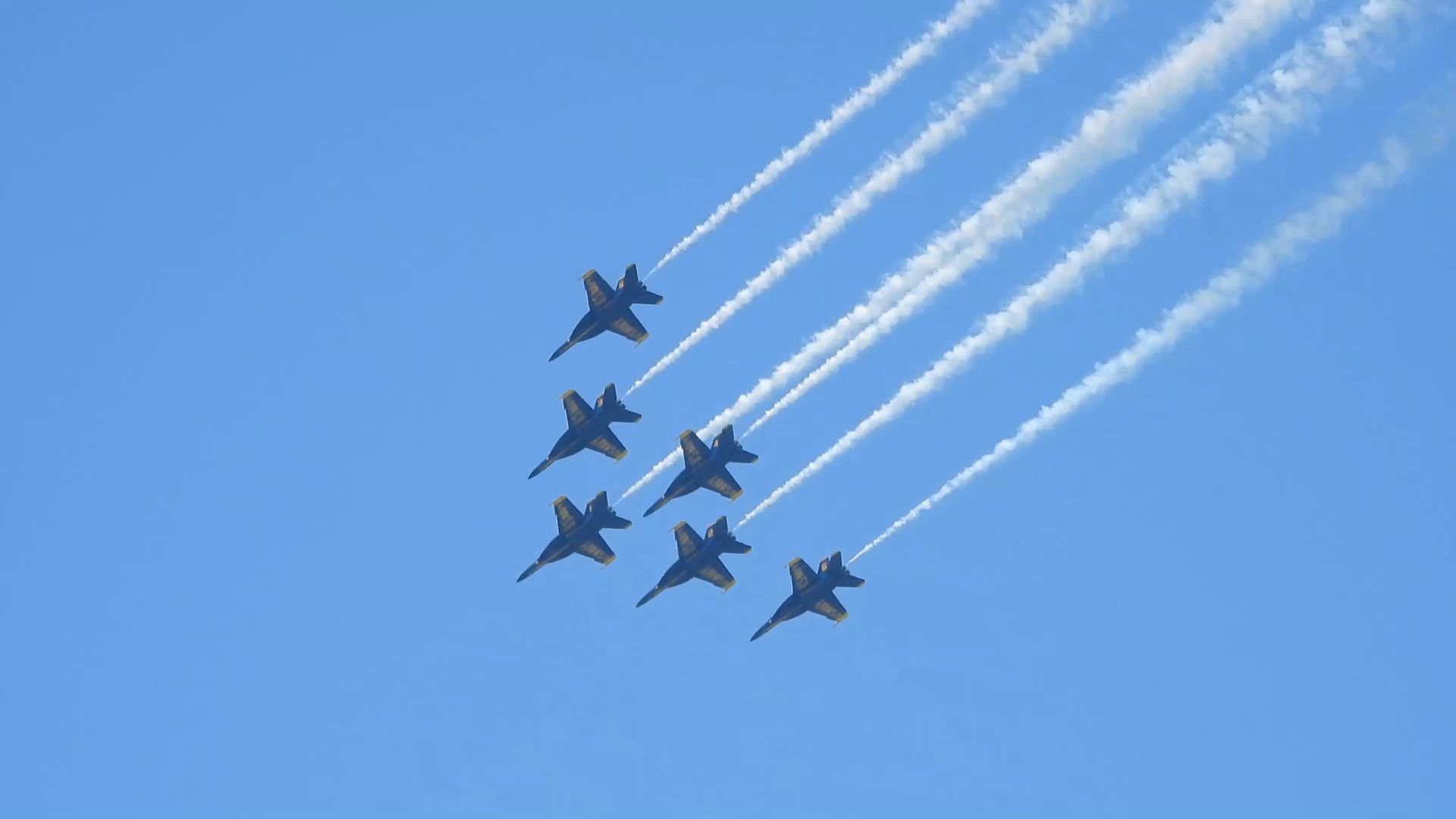 Six jets fly in close formation, trailing long parallel white smoke lines across a blue sky