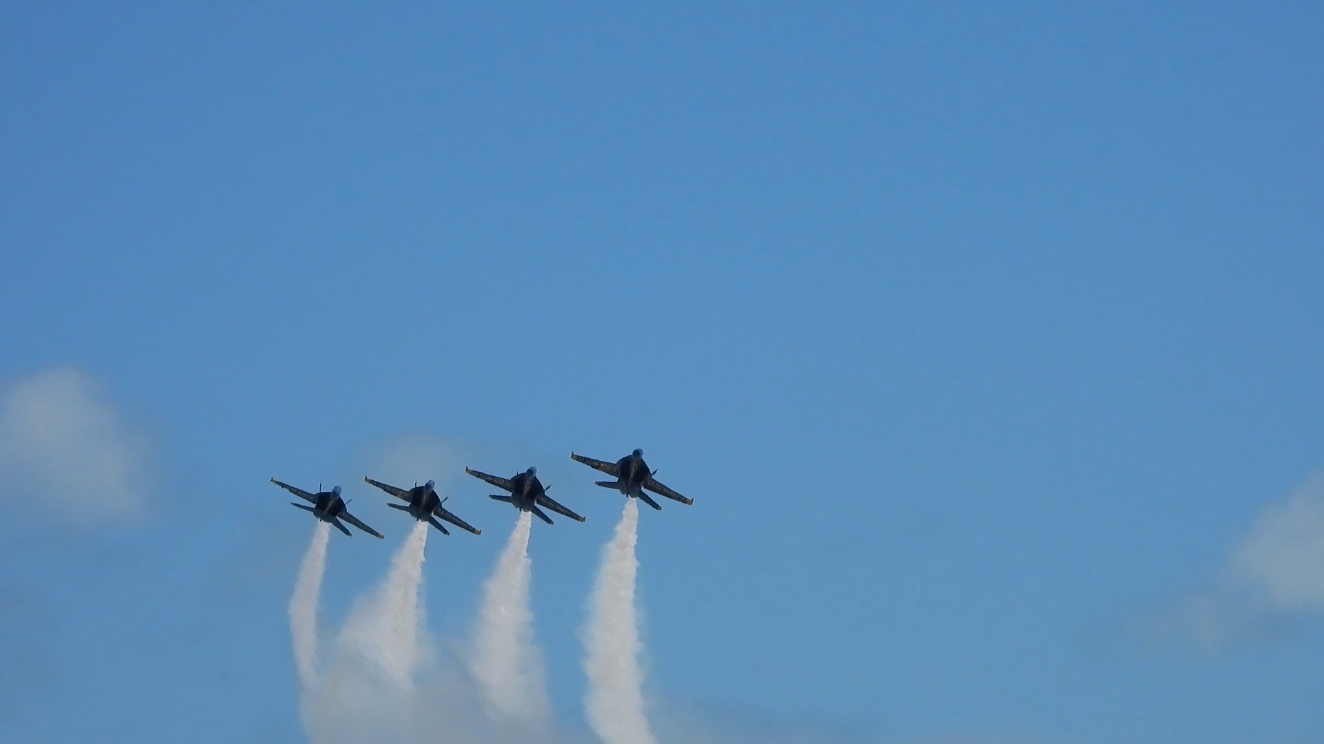 Four jets fly side by side in a line, white smoke billowing beneath them against a blue sky with clouds