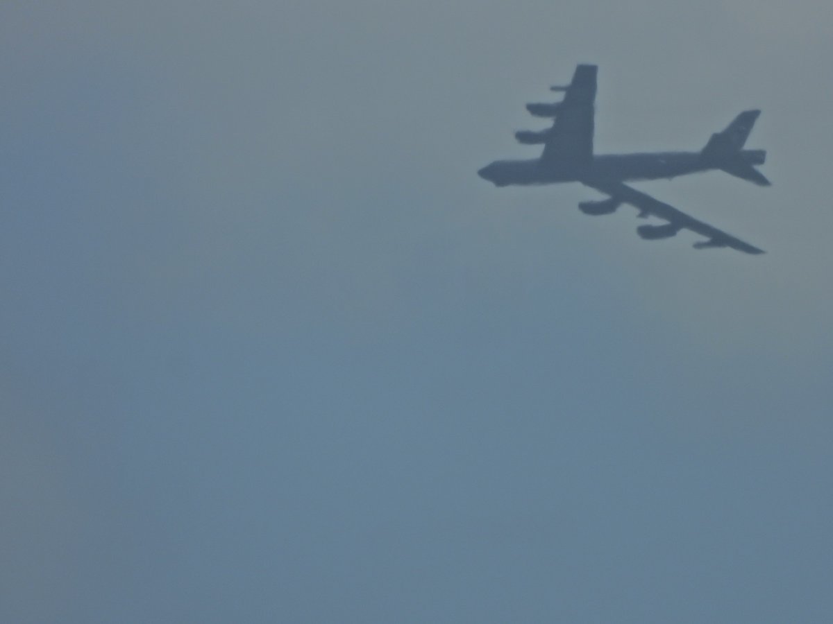 A large eight-engine bomber seen from below against a hazy gray sky