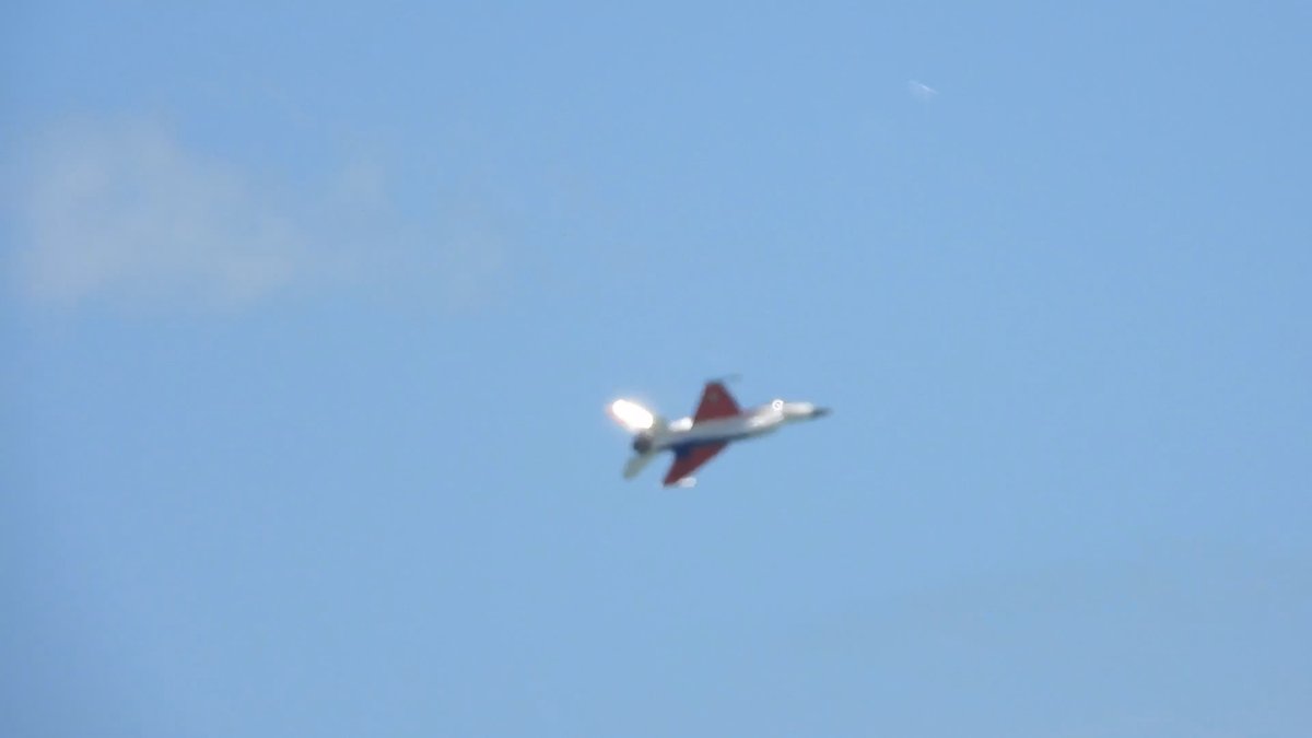 A gray fighter jet with a red-and-white tail flies against a pale blue sky