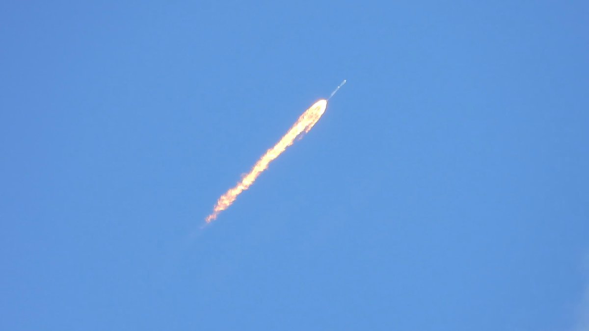 A bright orange rocket engine plume streaks diagonally upward through a clear blue sky