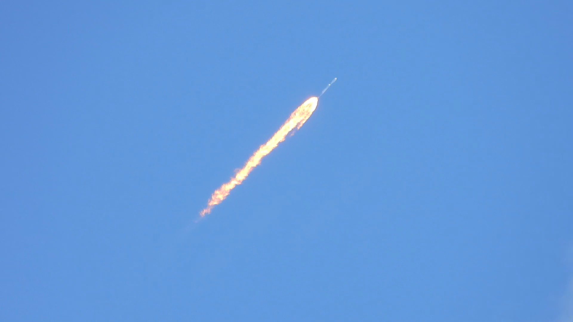 A bright orange rocket engine plume streaks diagonally upward through a clear blue sky